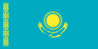 The flag of Kazakhstan has a turquoise field, at the center of which is a gold sun with thirty-two rays above a soaring golden steppe eagle. A thin vertical band displays a national ornamental pattern — koshkar-muiz — in gold near the hoist end.